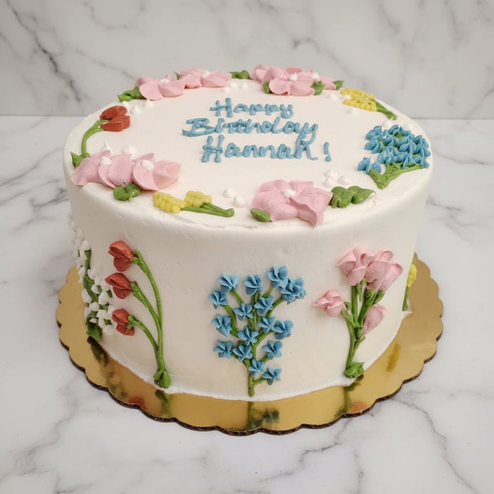 This classic buttercream cake features a smooth white buttercream finish adorned with hand-piped floral details inspired by a garden in bloom. Delicate flowers and leafy accents wrap the cake in soft color and texture, creating a fresh, cheerful design.