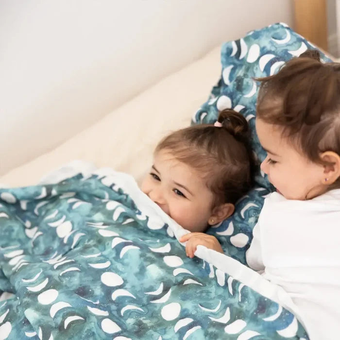 Two children lying under a blue patterned blanket on a bed.