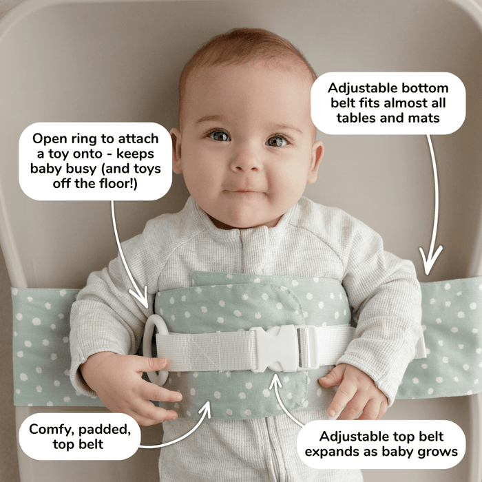 Baby lies on a change mat with an Oatmeal Afternoon WriggleBum nappy change harness wrapped around them. 
Four white bubbles highlight the key features of the harness: 
- Open ring to attach a toy onto - keeps baby busy (and toys off the floor!)
- Adjustable bottom belt fits almost all tables and mats 
- Adjustable top belt expands as baby grows
- Comfy, padded top belt